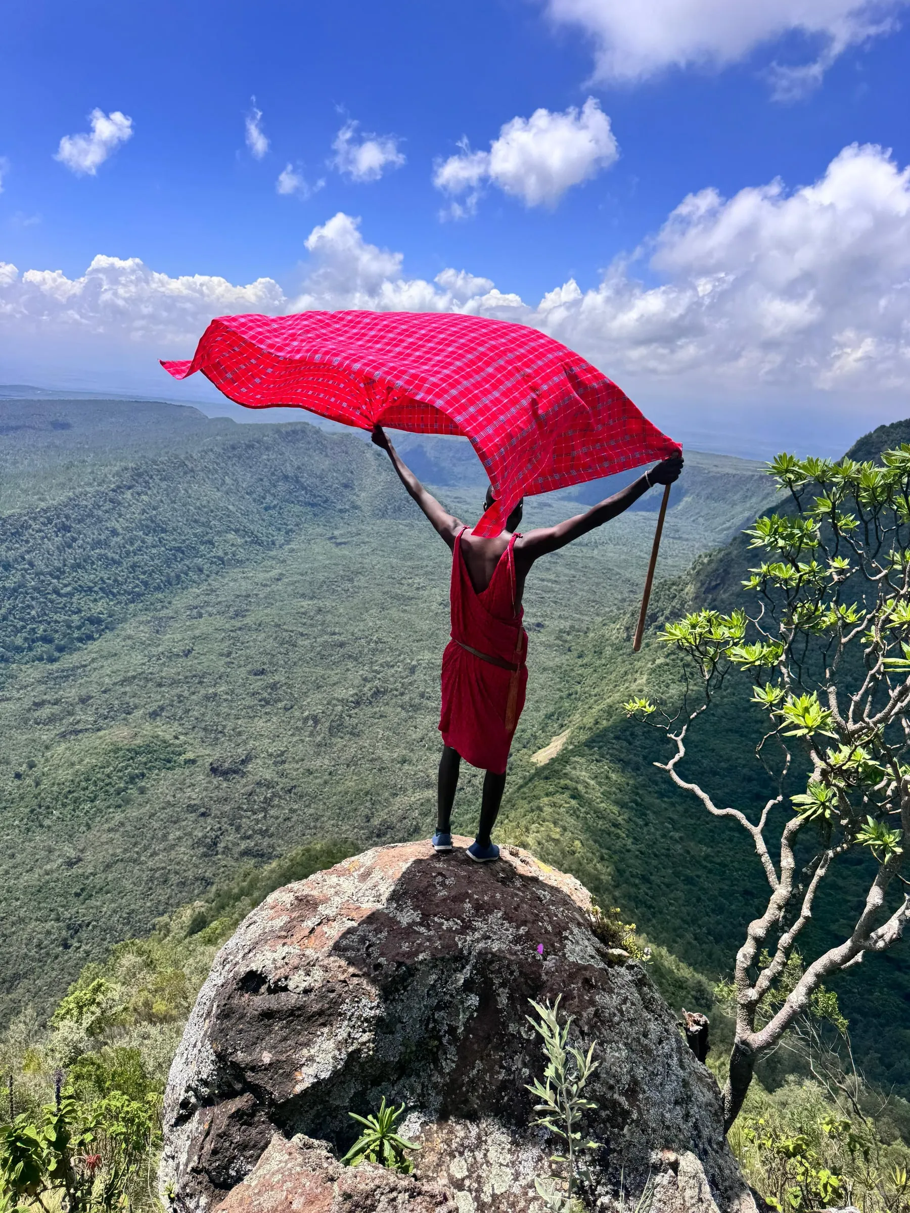 A Maasai guide in a red shuka on a rock above the crater, the cloth lifted by the wind